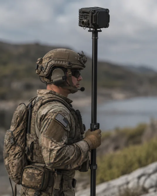 A soldier in helmet, headset, and multicam holding a mast-mounted QNV sensor head, coastal terrain behind.